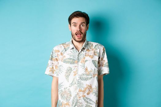 Summer holiday. Surprised guy in hawaiian shirt gasping amazed, checking out vacation offer, standing on blue background.