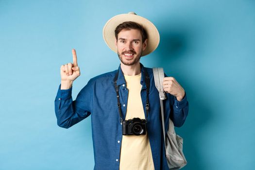 Handsome happy tourist in summer hat, holding backpack and camera, pointing finger up at logo, recommending travel agency or place on vacation, blue background.