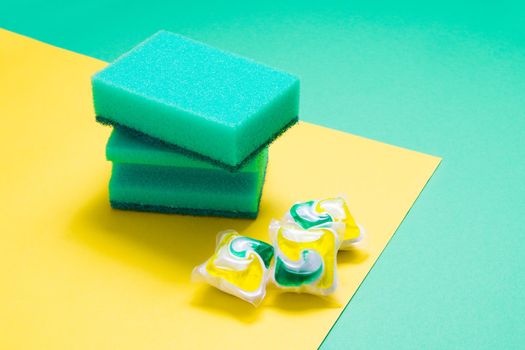 two green sponges for washing dishes and three capsules for dishwasher on a yellow and green background, copy space, blank for design
