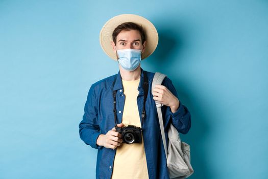 Covid-19 and travelling concept. Young guy tourist in medical mask and summer hat travel abroad during coronavirus pandemic, taking pictures on vacation, blue background.