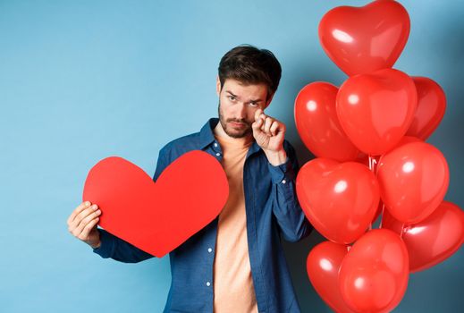 Sad and heartbroken man crying, wiping tears, standing with red heart and balloons, breakup on Valentines day, blue background.