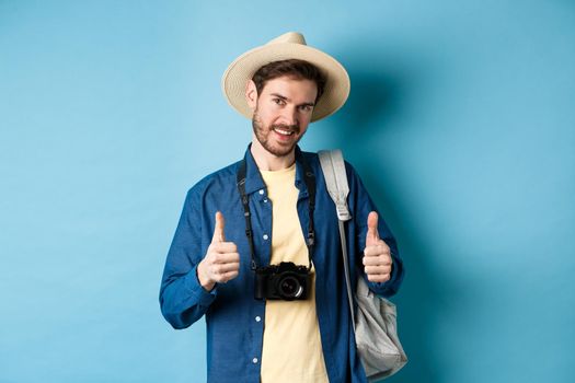 Handsome caucasian tourist in straw hat, holding backpack and photo camera, showing thumbs up, recommending travel agency or hotel resort, blue background.
