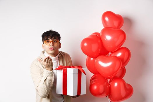 Valentines day and romance concept. Romantic modern man holding special gift for lover and sending air kiss at camera, standing near hearts balloons, white background.