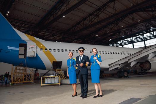 Full length shot of pilot in uniform and aviator sunglasses standing together with two air stewardesses in blue uniform in front of big passenger airplane in airport hangar. Aircraft, occupation