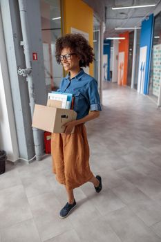 Full-length photo of smiling woman walking down the hallway in the office and carrying a box of documents