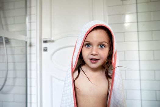 Cute kid with wet hair and in bathrobe standing in bathroom after taking shower and looking at camera