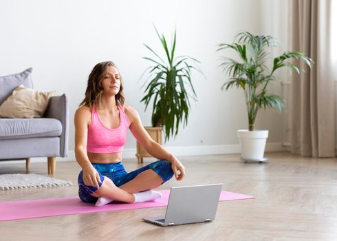 A young woman is meditating using a tutorial on the internet. She sits with closed eyes in front of a laptop monitor. Online workout during quarantine.