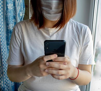 Medical mask on the face of an unknown girl, hands and mobile phone.The house near the window.
