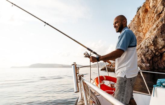 Young african american man standing with fishing rod on a sailboat fishing in open sea on sunset, close up