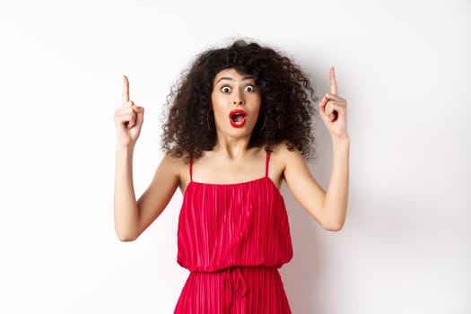 Impressed young woman with curly hair, wearing red dress, gasping and saying wow, pointing fingers up at logo, standing over white background.