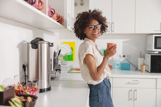 Pretty young woman holding coffee and posing on office kitchen while looking at camera
