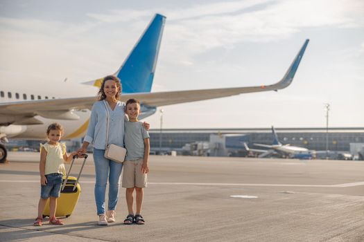 Full length shot of happy mother with two little kids smiling at camera while standing in front of big airplane in the daytime. Family, vacation, traveling concept