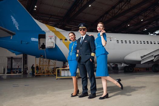 Full length shot of pilot in uniform and aviator sunglasses standing together with two air stewardesses in blue uniform in front of big passenger airplane in airport hangar. Aircraft, occupation