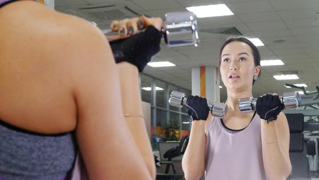 Training. A young motivated woman pumps her hand muscles with a dumbbells while looking in the mirror. Mid shot