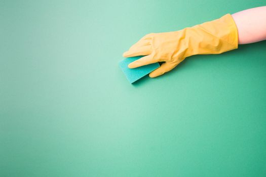female hand in a rubber glove holds a green paralonne sponge for cleaning and washing dishes, green background copy space, company cleaning and spring cleaning concept