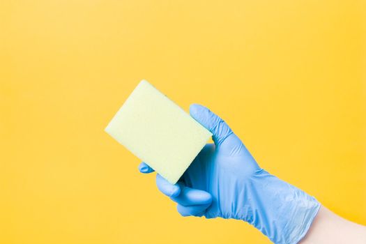 a hand in a blue disposable rubber glove holds a yellow paralon sponge, yellow background copy space, cleaning concept, home care, spring cleaning
