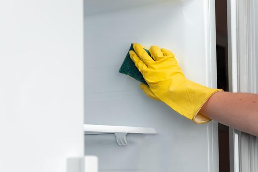 Hand of a woman in yellow glove cleaning the fridge shelf with a sponge