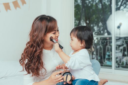 Innocent little daughter applying powder on her mom's face while sitting on bed at home