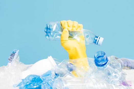 a hand in a yellow rubber glove squeezes a crumpled plastic bottle on a blue background, a hand in a pile of plastic debris, an environmental clogging with plastic