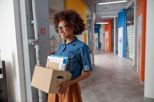 Smiling pretty woman in glasses carrying a box of documents down the hallway in the office, copy space