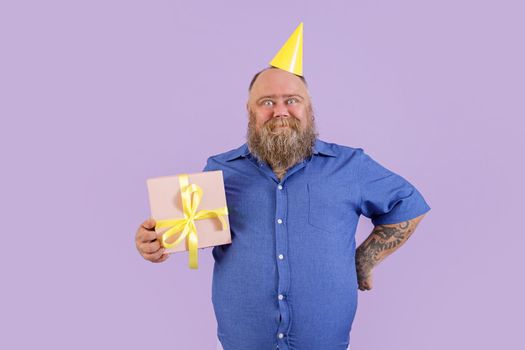 Positive mature man with overweight in tight blue shirt and yellow party hat holds gift box standing on purple background in studio