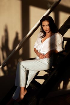 Young confident female wearing white shirt and pants sitting on wooden stairway and looking at camera