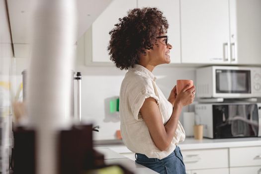 Side view of smiling multiethnic woman holding cup of coffee while standing at office kitchen