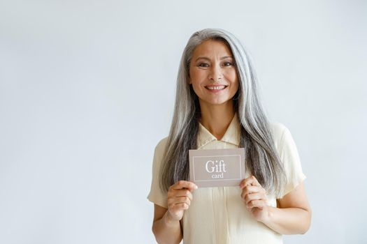 Smiling grey haired Asian woman holds gift card standing on light background in studio, space for text. Shopping certificate