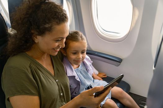 Happy mother holding mobile phone and looking at the screen while traveling with her daughter on an airplane