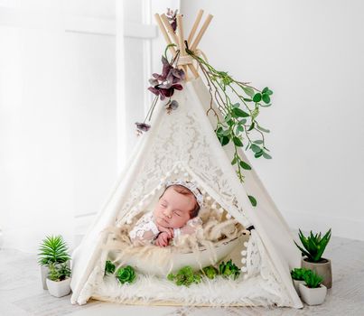 Adorable newborn baby girl wearing beautiful dress and wreath lying in hut wigwam with plant decoration holding hands under her cheeks in studio. Cute infant child napping on fur