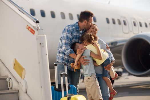Family of four kissing each other while going on a trip, standing in front of big airplane outdoors. People, traveling, vacation concept
