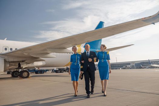 Full length shot of professional pilot walking together with two cheerful stewardesses in bright blue uniform in front of an airplane on a sunny day after landing. Aircraft, aircrew concept