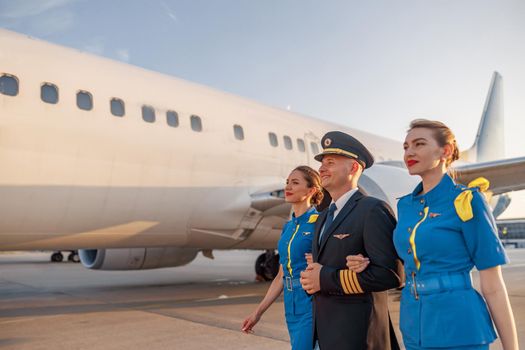 Excited male pilot walking together with two flight attendants in blue uniform in front of an airplane in terminal at sunset. Aircraft, aircrew concept