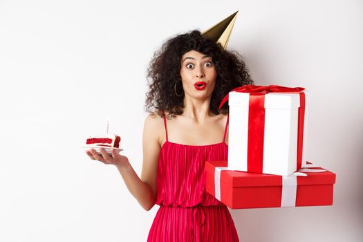 Funny lady in red dress and party hat, celebrating birthday, holding b-day presents and cake with candle, looking amused at camera, standing over white background.
