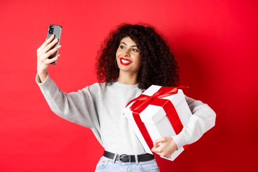Happy young woman taking selfie with her valentines day gift, holding present and photographing on smartphone, posing on red background.