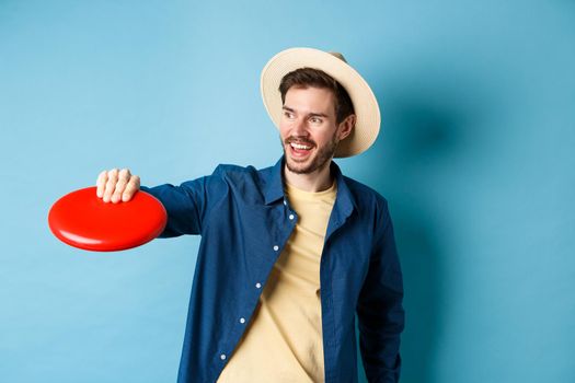 Happy smiling guy catching frisbee while playing with friends on summer vacation, standing in straw hat on blue background.