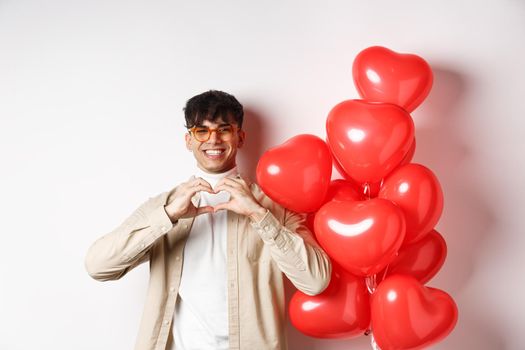 Valentines day and romance concept. Handsome young man standing near red hearts balloons and showing heart gesture, being in love and smiling, white background.