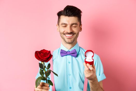 Valentines day. Cute boyfriend making wedding proposal, showing engagement ring in small box and red rose, express love, standing over pink background.