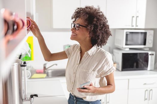 Smiling young woman holding smartphone while going to make coffee on office kitchen