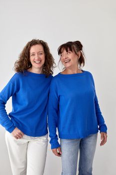 Glad loving mature mother and adult daughter wearing similar blue sweaters on gray background in studio