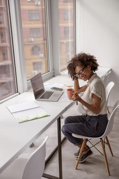 Happy Afro American female worker enjoying coffee while sitting at desk near the window