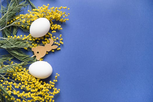Easter background, white eggs and a wooden hare on a blue background, decorated with Mimosa flowers, flatlay, top view, empty space for text. happy Easter.