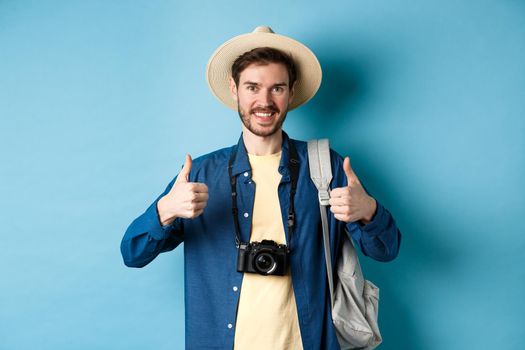 Cheerful handsome man recommending summer vacation place, showing thumbs up and smiling in approval. Tourist leave positive feedback, standing with camera and backpack on blue background.