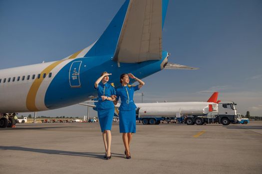 Full length shot of two cheerful air stewardesses in bright blue uniform walking outdoors in front of passenger aircraft on a sunny day. Occupation, aircrew concept