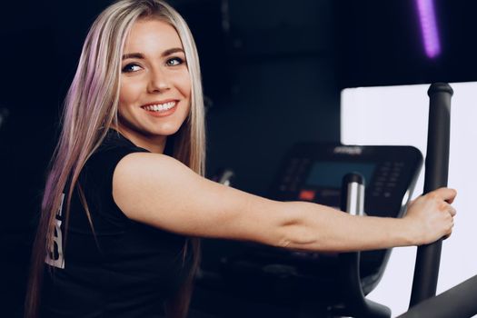 Attractive young blonde woman exercising on cardio training apparatus in dark gym