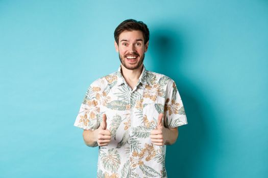 Summer holiday. Excited smiling tourist in hawaiian shirt show thumbs-up, praising hotel or vacation place, standing on blue background.