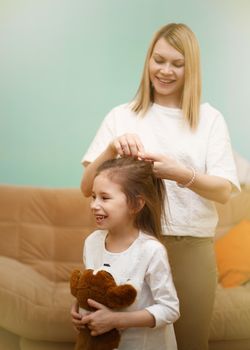 Mother is braiding her young daughter's hair standing in the children room. Happy loving family.