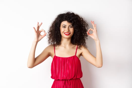 Beautiful woman in red dress, smiling and showing okay gestures, like something good, praising good job, standing over white background.