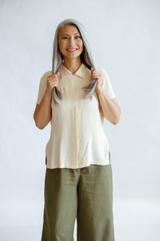 Smiling middle aged Asian woman in casual clothes holds long healthy grey hair standing on light background in studio. Mature beauty lifestyle