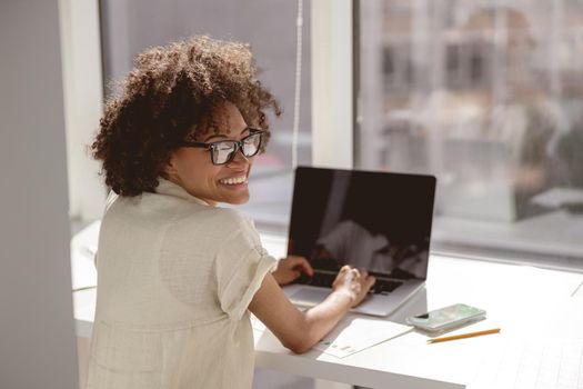 Happy Afro American lady in glasses typing on laptop while sitting at her workplace. Copy space